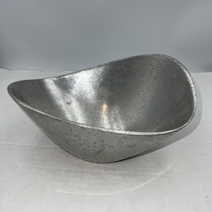 May include: A small, silver-colored bowl with a unique, elongated shape. The bowl has a textured surface and a slightly curved rim. The bowl is made of metal and appears to be vintage.