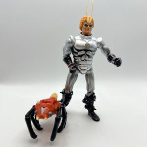 May include: A plastic action figure with orange hair, blue eyes, and a silver chest plate, wearing black boots and wrist guards. A small, orange, spider-like creature with black legs and white fangs stands on the ground next to the figure.