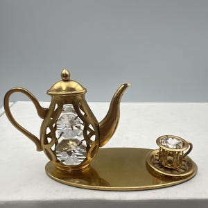 May include: A gold-colored decorative tea set featuring a teapot with a crystal insert and a teacup with a saucer. The teapot and teacup have intricate openwork designs. The set is displayed on a gold-colored oval tray.