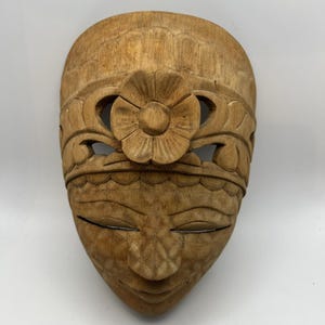 Op de afbeelding: Een lichtbruin houten wandmasker met een uitgesneden bloemmotief op het voorhoofd, gebogen wenkbrauwen, gesloten ogen en een geometrisch patroon op de neus en wangen. Het masker heeft een gladde, natuurlijke houtnerf en een afgeronde bovenrand.