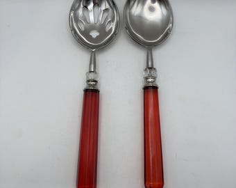 Può includere: Una coppia di cucchiai da portata color argento con coppe in metallo lucido e manici sfaccettati in rosso traslucido. Un cucchiaio presenta un design a fessura con aperture multiple, mentre l'altro è un cucchiaio solido e liscio.