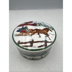 Può includere: Un portagioie rotondo in ceramica bianca con un coperchio raffigurante una scena invernale. Il coperchio raffigura una slitta trainata da cavalli, un fienile rosso e alberi innevati. La scatola ha un bordo verde e un disegno di recinzione marrone.
