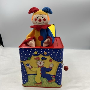 Pode incluir: Um brinquedo Jack-in-the-box vintage. A caixa de lata vermelha e azul apresenta uma ilustração de palhaço de circo e uma manivela. Uma boneca de palhaço de pelúcia com um chapéu amarelo, roupa vermelha e azul e bochechas rosadas sai da caixa.