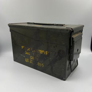 May include: An olive drab metal ammunition box with a hinged lid and a latch closure. Yellow text on the side reads "100 CAL. CARTRIDGES BALL M1917 CARTON LOT RA 895". The box has a handle on top and shows signs of wear.