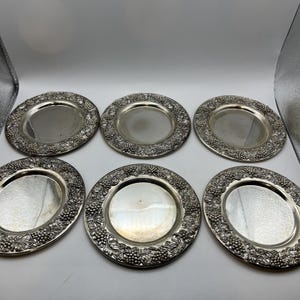 May include: A set of six circular silver-toned metal plates, each featuring a smooth, reflective center and a wide, raised border decorated with a detailed, embossed pattern of grape clusters and vine leaves encircling the entire outer rim.