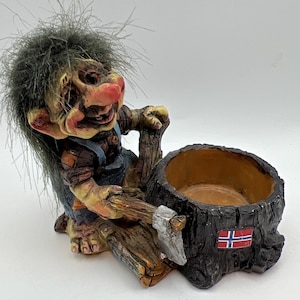 May include: A whimsical troll figurine with gray hair, a large red nose, and blue overalls, holding an axe. The troll is positioned next to a tree stump planter with a Norwegian flag. The planter has a brown interior.