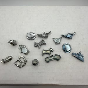 May include: A collection of fourteen silver-toned metal game tokens featuring various shapes including a top hat, a shoe, a hand, a dog, a pretzel, a race car, a ship, a thimble, and a set of teeth, all arranged on a plain white surface.