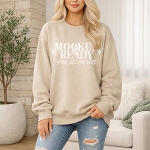 Puede incluir: Una sudadera beige con el texto "MOCKEL READY your DESIGN HERE" en blanco. La sudadera es de cuello redondo y se usa con vaqueros azules. La imagen es una maqueta para la colocación de diseños.