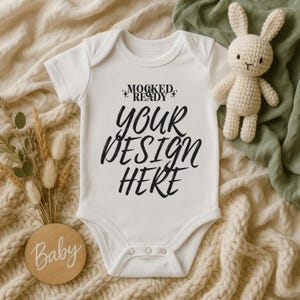 May include: A white baby onesie with the text "YOUR DESIGN HERE" in black script. A small crocheted bunny and a wooden disc with the word "Baby" are also in the image. The onesie is on a cream-colored blanket.