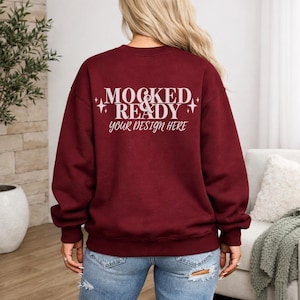 May include: A maroon sweatshirt with the text "MOCKED & READY your design here" in white script. The sweatshirt is a solid color and has a crew neck. The model is wearing blue jeans.