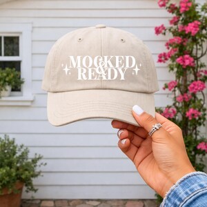 May include: A beige baseball cap with the text "MOCKED & READY" in white. The cap is held up by a hand with a ring. The background features a white house and pink flowers.