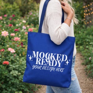 May include: A royal blue tote bag with white text that reads "MOCKED & READY your design HERE". The bag has long straps and is being held by a person. The background is a garden with flowers.