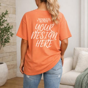May include: Orange t-shirt with the text "YOUR DESIGN HERE" in white script. The shirt is a solid color and has short sleeves. The model is wearing blue jeans.