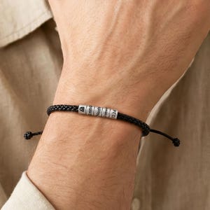 May include: A black braided cord bracelet featuring a cylindrical silver-toned metal bead engraved with text. The bracelet has adjustable knotted ends and is worn on the wrist, showcasing a minimalist design with a metallic focal point.