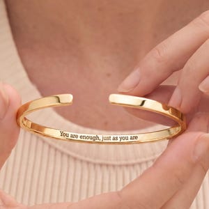 May include: A gold-toned open cuff bracelet with a polished finish. The interior surface of the metal band features the engraved text, "You are enough, just as you are," written in a clear, dark, sans-serif font centred along the inner curve.