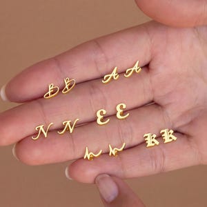 May include: A selection of gold-coloured initial stud earrings in various cursive fonts. The letters include A, B, E, H, K, and N. The earrings are presented against a light brown backdrop.