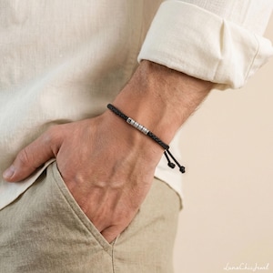May include: A black cord bracelet featuring a single cylindrical silver-toned metal bead with engraved markings. The bracelet is secured with a simple knot closure, and the cord has a braided texture that wraps around the wrist.