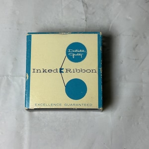 Może przedstawiać: Kwadratowe kartonowe pudełko na taśmę barwiącą z kremowym przodem, niebieskimi obrzeżami i niebieskim tekstem. Pudełko zawiera napis Inked Ribbon, niebieski ząbkowany okrąg z tekstem Distinctive Quality oraz napis Excellence Guaranteed.