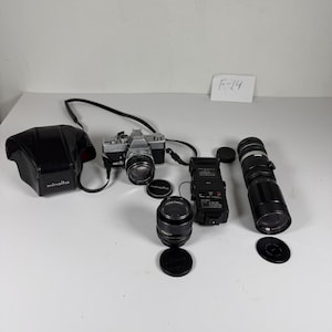 Puede incluir: Una cámara de película Minolta SRT 101 plateada y negra con lente, un estuche de cuero negro, un flash electrónico negro, un lente secundario, un teleobjetivo largo y tres tapas de lente circulares. Se incluye una tarjeta blanca con F-14.