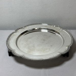 Könnte beinhalten: Ein rundes silberfarbenes Serviertablett mit einer glatten, reflektierenden Mitte und einem dekorativen, gewellten Rand, der ein gedrehtes, seilartiges Muster aufweist. Das Tablett hat eine polierte metallische Oberfläche und einen breiten Rand.