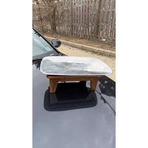 Könnte beinhalten: Ein rechteckiges, silberfarbenes Serviertablett mit abgerundeten Ecken steht auf einem kleinen Holzständer. Der Ständer hat vier kurze Beine und eine natürliche Holzoberfläche. Das Tablett reflektiert die Umgebung.