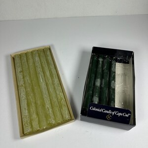 May include: Two rectangular boxes containing taper candles. The left box holds six light green candles. The right black box contains three dark green candles and is labelled Colonial Candle of Cape Cod, with an empty slot for a fourth candle.