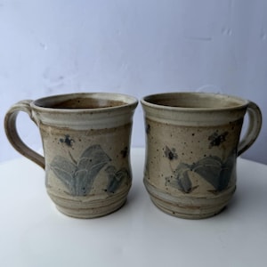 Puede incluir: Dos tazas de cerámica con acabado beige moteado y un patrón floral azul pintado a mano. Cada taza cuenta con un asa resistente, un borde ancho y una base ligeramente cónica. La superficie tiene una apariencia terrosa y texturizada.