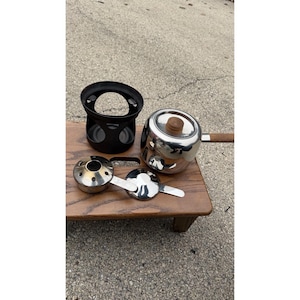 Puede incluir: Un juego de fondue de acero inoxidable con una olla con mango y tapa de madera, un quemador de metal negro y dos soportes de combustible de acero inoxidable. Los artículos están colocados sobre una pequeña mesa de madera.