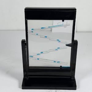 Puede incluir: Un juguete de movimiento líquido de plástico negro con un panel rectangular transparente y pistas internas en zigzag. Gotas de líquido azul brillante descienden por los canales escalonados dentro del marco sobre base.
