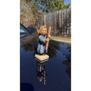 Op de afbeelding: Een klein beeldje van een kinderengel met een roze kaars met een brandende oranje vlam. De engel draagt een blauwe jurk met gouden sterren en een donkerblauwe mantel. Het beeldje staat op een vierkante basis.