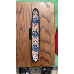 May include: A long, narrow ceramic dish with a blue rim and a floral pattern of blue and peach flowers on a white background. The dish is resting on a wooden surface.