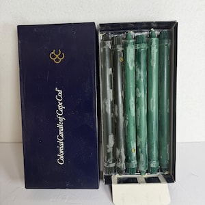 May include: A set of six dark green taper candles housed in a rectangular navy blue box with the text Colonial Candle of Cape Cod printed in white. The candles feature a textured, weathered finish and are arranged vertically inside the open box.