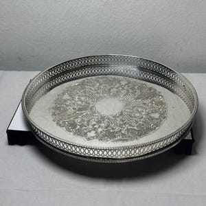 May include: A round silver-colored metal tray featuring an intricate embossed floral and scrollwork pattern in the center. The tray has a raised, openwork lattice border around the entire circumference, creating a decorative gallery edge.