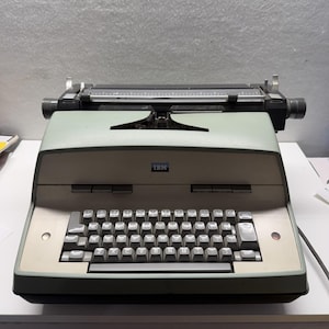 May include: A vintage IBM electric typewriter featuring a sage green casing, a QWERTY keyboard with grey keys, and a black carriage assembly. The IBM logo is centred on the front panel, with a paper feed mechanism and knobs visible at the top.