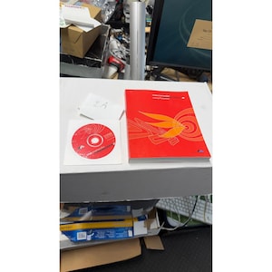 Könnte beinhalten: Ein rotes Buch und eine CD-ROM mit dem Text "Macromedia Fireworks" werden auf einer weißen Oberfläche angezeigt. Das Buch hat ein stilisiertes orange-gelbes Design. Eine kleine weiße Karte mit dem handschriftlichen Text "2A" ist ebenfalls vorhanden.