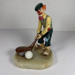 May include: A clown figurine wearing a dark green cap, yellow shirt, pink bow tie, and dark green pants, holding a brown golf club over a white golf ball. The figure stands on a textured, light-colored base with sandy patches under the feet and ball.