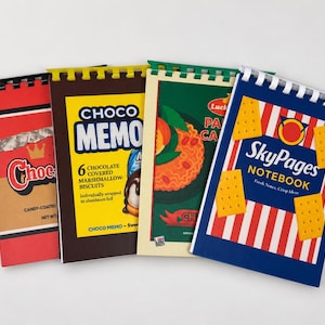 May include: Four spiral-bound notebooks with covers designed to resemble food packaging. The designs include a red Choco wrapper, a yellow Choco Memo box, a green pasta package, and a blue SkyPages notebook with cracker illustrations.
