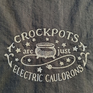 May include: Black fabric with silver embroidered text and a cauldron design. The text reads "CROCKPOTS are just ELECTRIC CAULDRONS", surrounded by stars and decorative flourishes. The design is centered on the fabric.