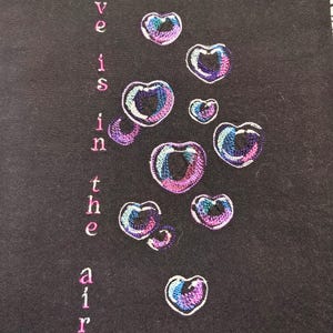 May include: Black fabric with embroidered heart-shaped bubbles in shades of purple, blue, and pink. The words "Love is in the air" are embroidered vertically in pink letters.