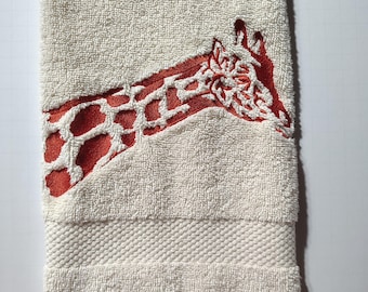 May include: Cream-colored towel with an embroidered giraffe design in red and white. The giraffe's head and neck are prominently displayed on the towel, which has a textured border and the text "moto g stylus - 2025" and "DMCsFindsAndDesigns".
