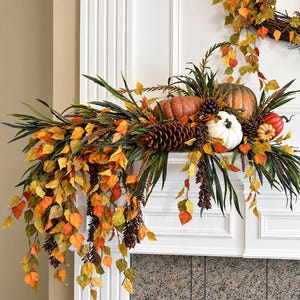 May include: A decorative autumn mantel swag featuring a mix of orange, yellow, and green faux leaves, brown pinecones, and several pumpkins in shades of orange, white, and green. The arrangement includes long, slender green foliage.