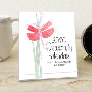 May include: A 2026 Dragonfly calendar with a watercolor illustration of a red dragonfly. The calendar is in a clear plastic stand. The text on the calendar reads "2026 Dragonfly calendar watercolor illustrations by bill semann."