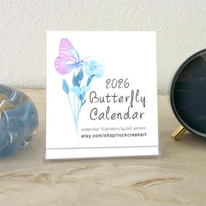 May include: A 2026 Butterfly Calendar featuring a watercolor illustration of a pink and blue butterfly with matching flowers. The calendar is displayed in a clear plastic stand on a light-colored surface. The text "watercolor illustrations by bill semann etsy.com/shop/rockcreekart" is also visible.