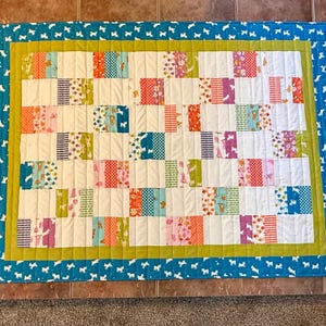 May include: A colorful patchwork quilt with a blue border featuring white dog silhouettes. The quilt's interior has a grid pattern of various patterned squares in shades of pink, orange, green, and blue, with white spaces in between.