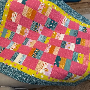 May include: A handmade quilt with a patchwork design. The quilt features large pink squares interspersed with smaller squares of various patterns and colors, including teal, yellow, and white. The border is teal with a white floral pattern.