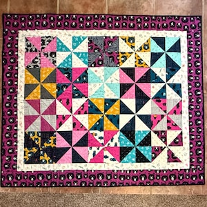 May include: A colorful quilt with a pinwheel design. The quilt features squares of pink, teal, yellow, and gray fabrics. The border is a dark purple with a repeating bear pattern. The quilt is a rectangle.