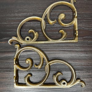 May include: Two matching gold-toned metal shelf brackets featuring an ornate, curved scrollwork design. Each bracket has a flat horizontal base and a vertical back plate with visible screw holes for mounting, showcasing a polished metallic finish.