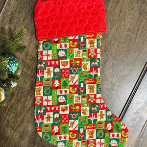 May include: A quilted Christmas stocking with a red cuff and a patterned body. The body features a patchwork design with festive images like gingerbread men, mice, and wrapped gifts in red, green, and white squares. A red loop is attached to the cuff.