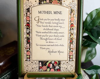 May include: A framed print with the text "Mother Mine" in elegant script. The green wooden frame surrounds a cream-colored paper with a poem and decorative floral accents in red, pink, and black. The print is displayed on a decorative stand.