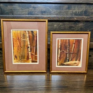 May include: Two framed watercolor paintings depicting a forest scene with white birch trees against a warm orange and brown background. Each piece is enclosed in a gold-toned frame with a wide brown mat and a thin white inner border.